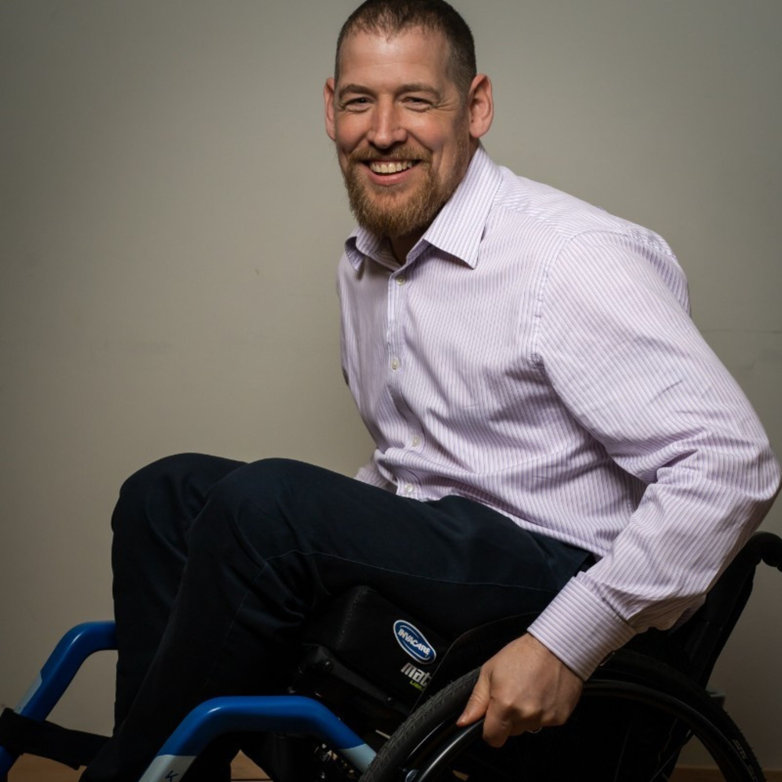 Jamie McAnsh, seated in a wheelchair, smiling warmly while wearing a light pink striped shirt and dark trousers, exuding confidence and positivity.